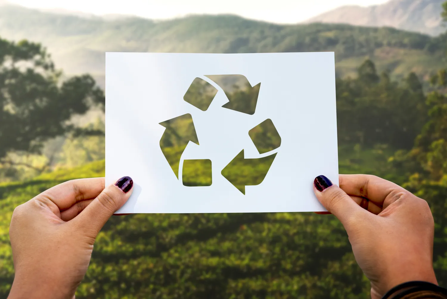 Zwei Hände halten ein Blatt Papier mit dem Recyclingsymbol vor einer grünen, hügeligen Landschaft.